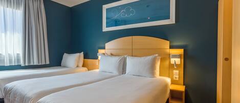 Quarto de hotel com duas camas, uma janela grande com cortinas, uma obra de arte na parede e uma mesa de cabeceira com abajur.