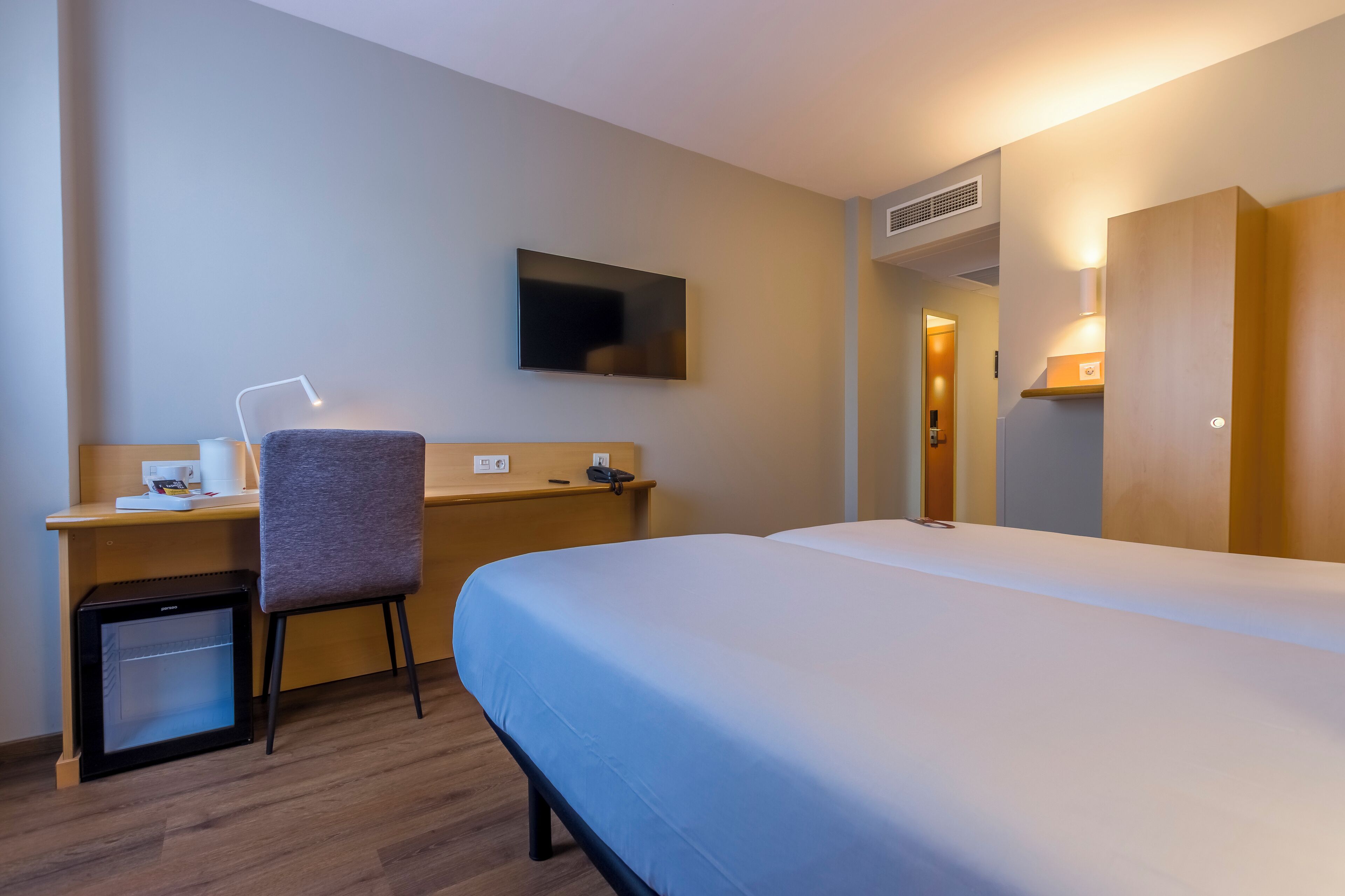 Una habitación de hotel con cama, escritorio, silla y un refrigerador pequeño.
