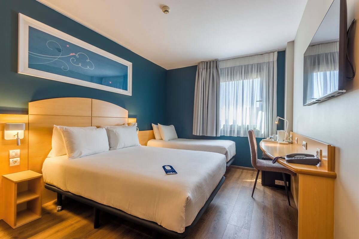 A hotel room with two beds, a desk, a chair, and a large window with curtains.