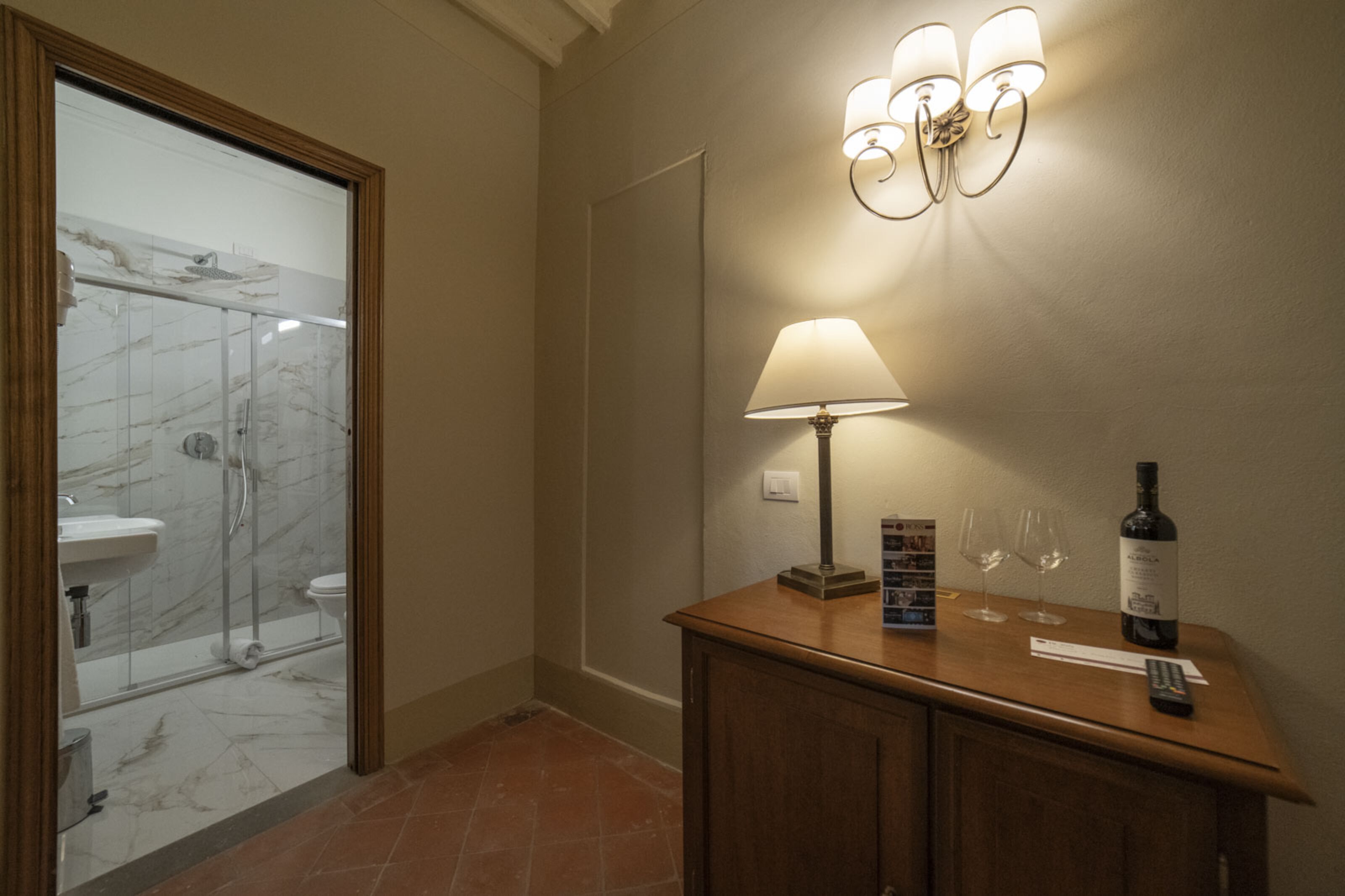 A hotel bathroom with a glass shower enclosure, a sink, and a vanity with a bottle and glasses.
