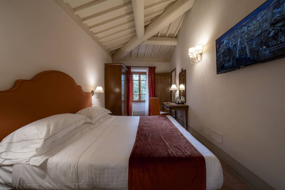A hotel room with a large bed, a wooden headboard, a desk with a chair, a mirror, and a painting on the wall.
