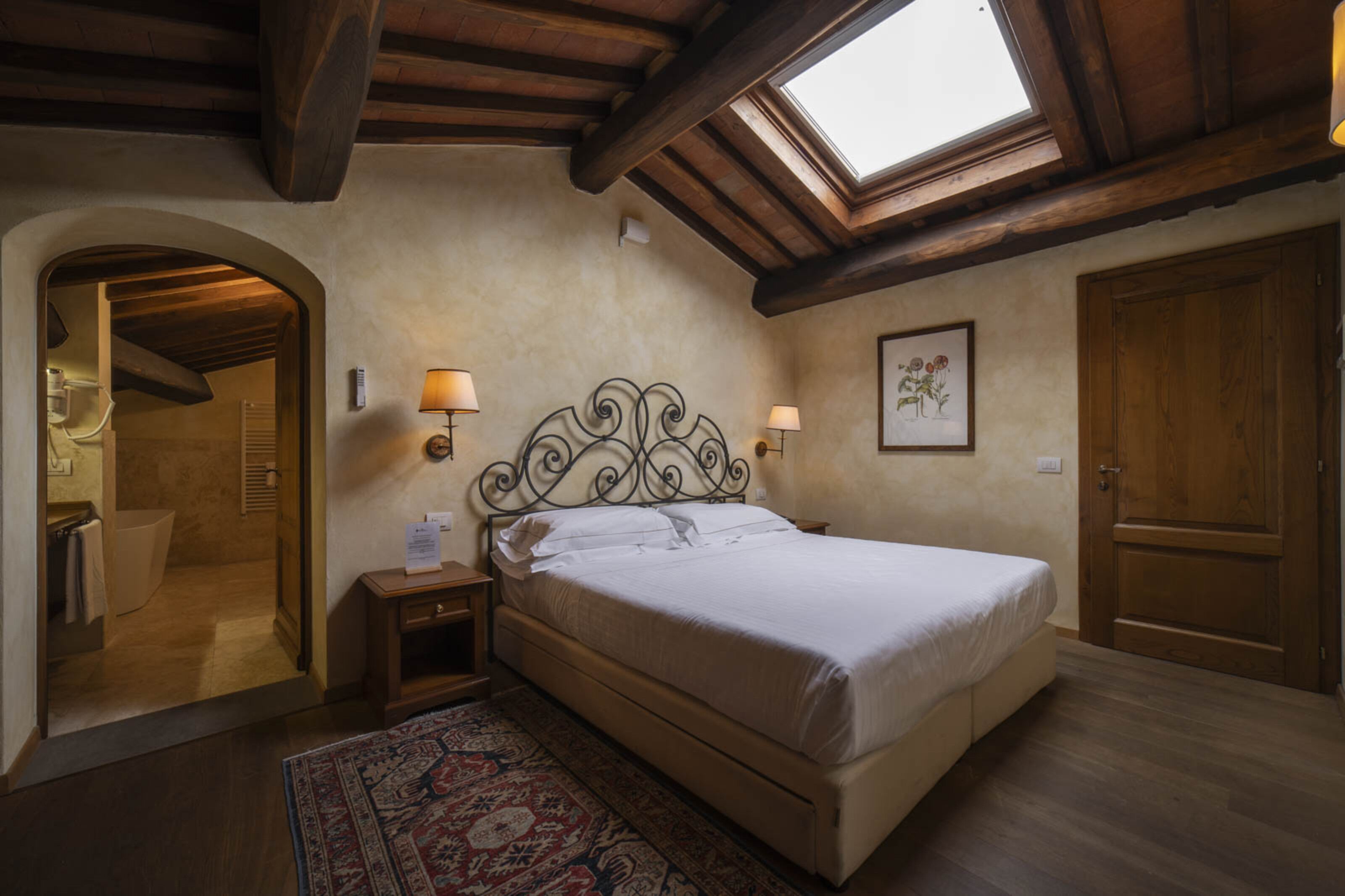 A bedroom with a skylight, a bed, a nightstand, a mirror, a door, and a wall-mounted lamp.