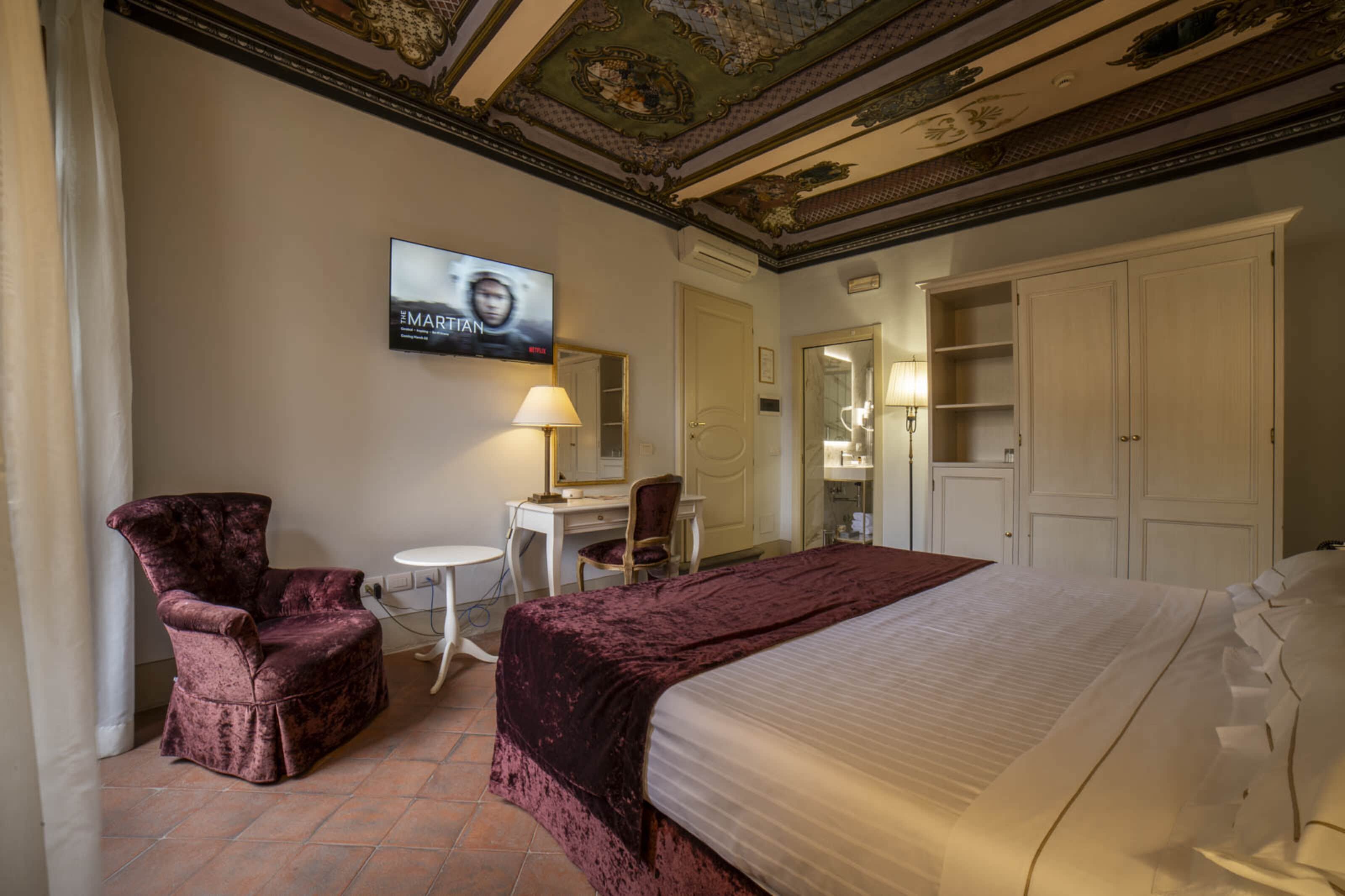 Ein Hotelzimmer mit einem Bett, einem Schreibtisch, einem Sessel und einem Fernseher, der einen Film zeigt.