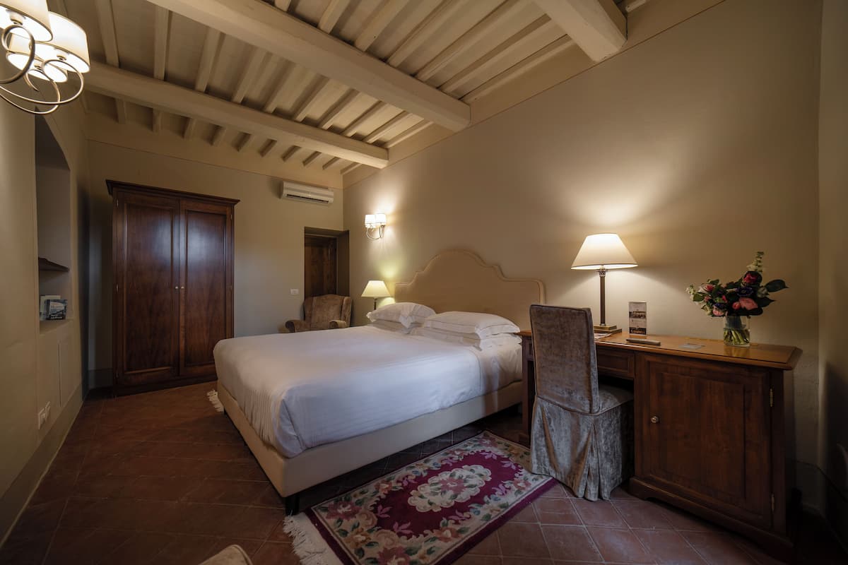 A bedroom with a large bed, a wooden wardrobe, a desk with a lamp, and a floral rug.