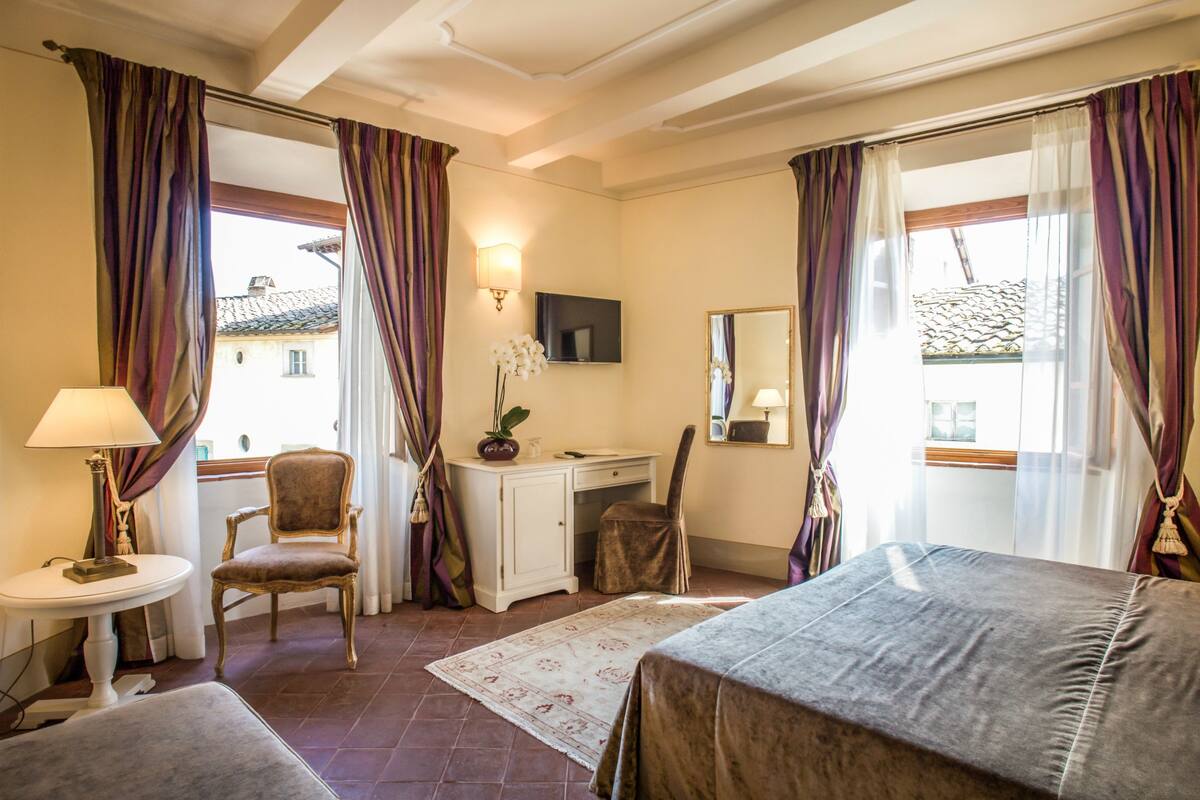 A hotel room with a bed, desk, chair, and a small table with a lamp.