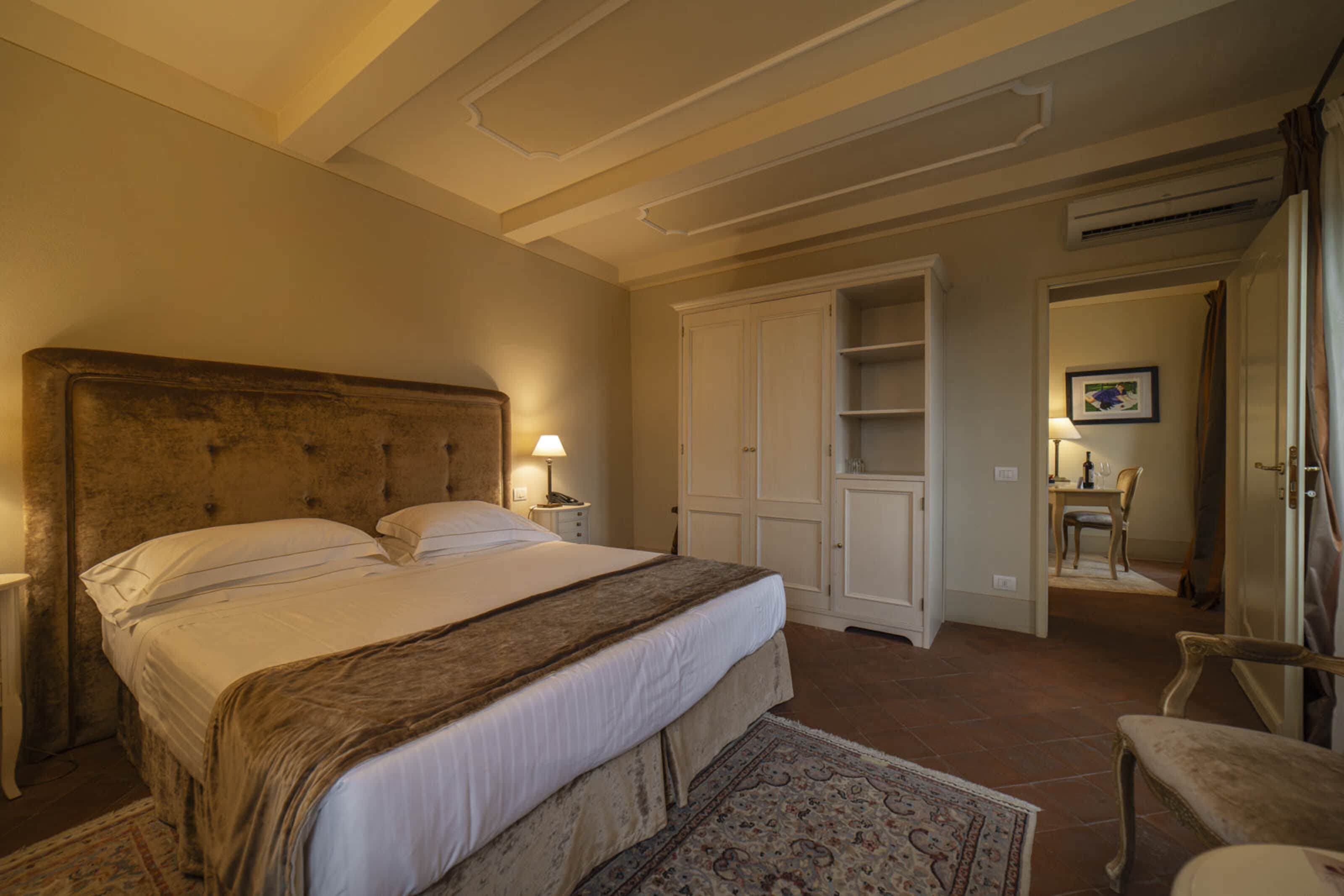 A bedroom with a large bed, a nightstand, a lamp, a wardrobe, and a small table with a chair.