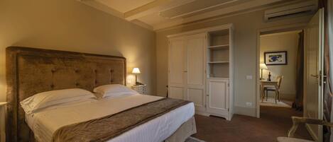 A bedroom with a large bed, a nightstand, a lamp, a wardrobe, and a small table with a chair.