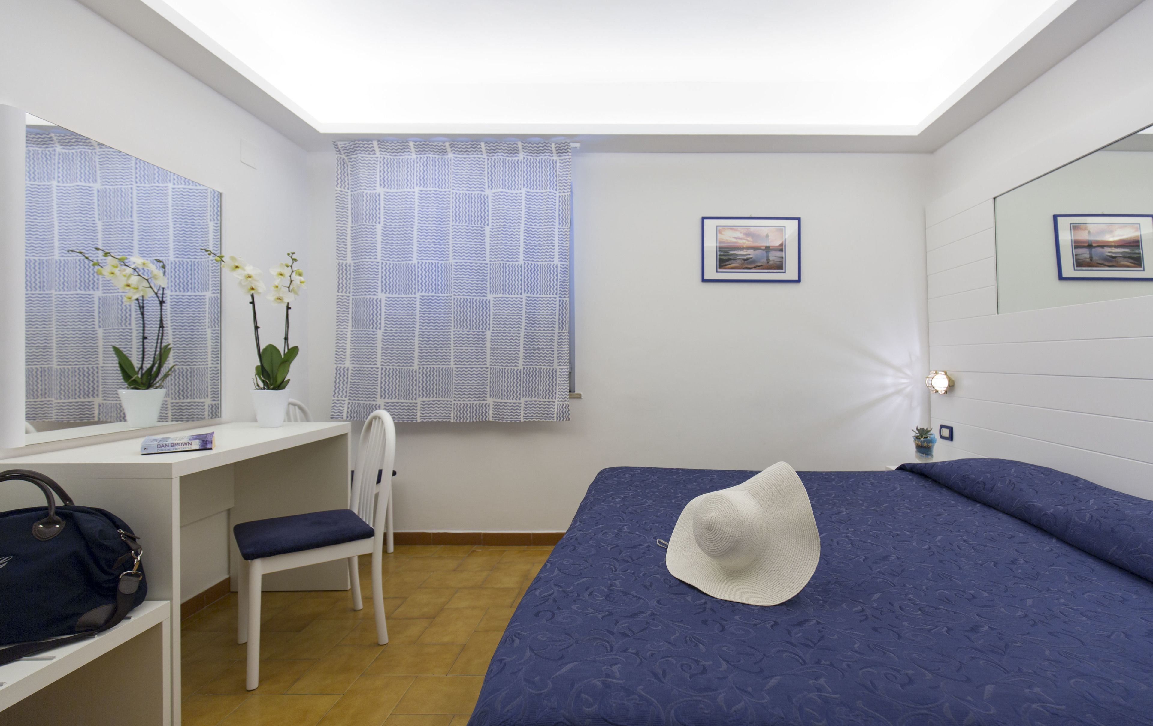 Habitación de hotel con una cama, un escritorio, una silla, una mesita con una planta y una ventana con cortinas estampadas.