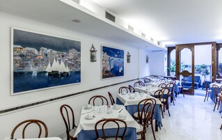 Un comedor con mesas dispuestas para el servicio, un cuadro grande de un paisaje urbano costero y vista al mar a través de un vano abierto.