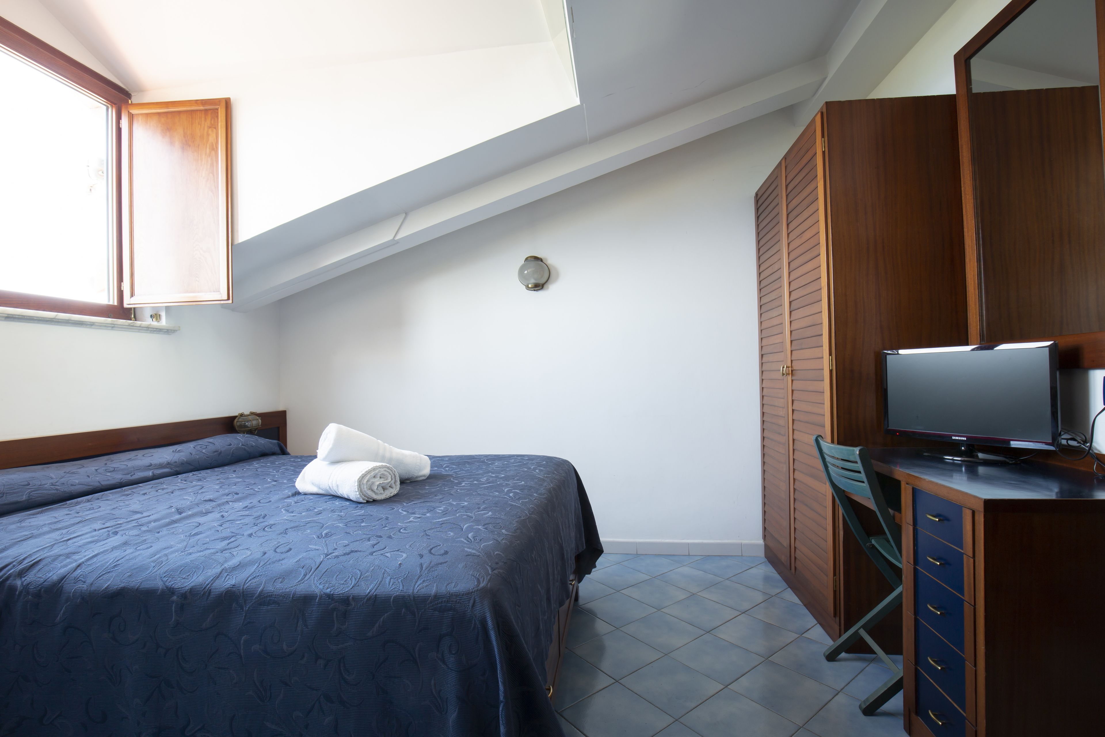 A bedroom with a bed, a desk with a computer, a window, and a wardrobe.
