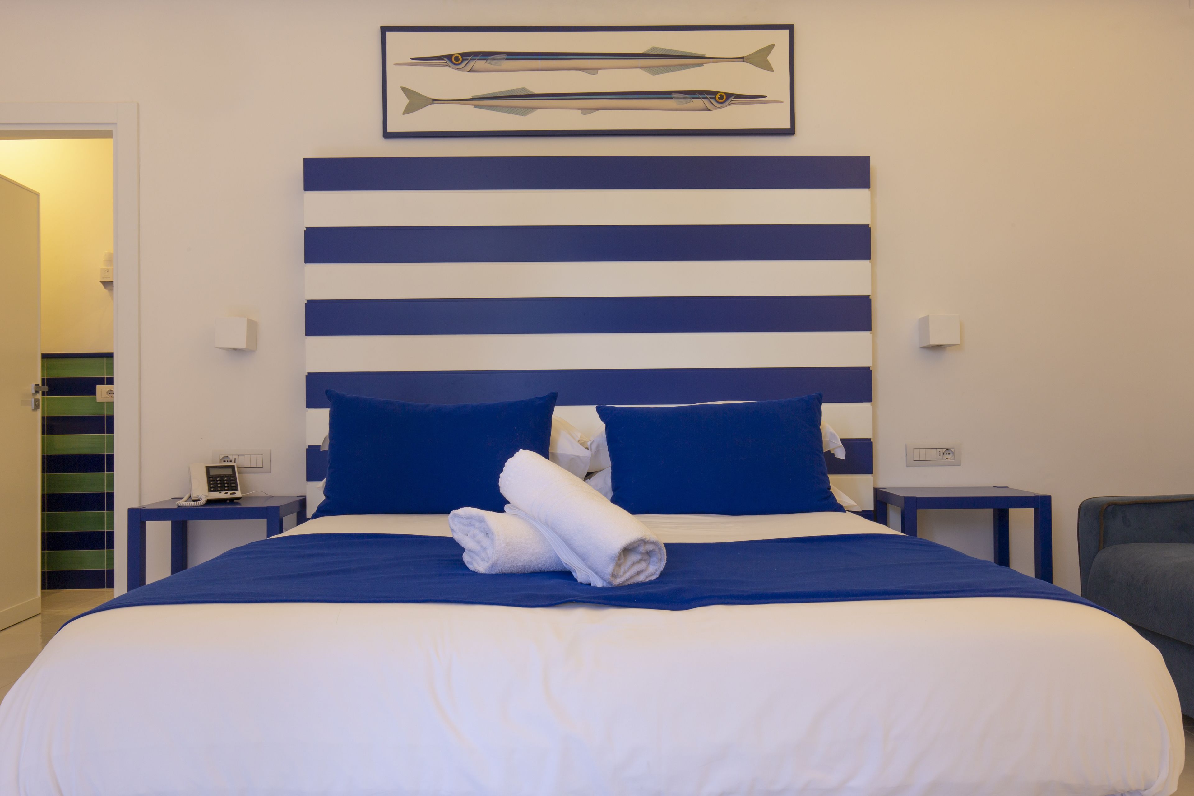 A hotel room with a large bed, blue and white striped headboard, two pillows, a framed picture of fish, a bedside table with a phone, and a chair.