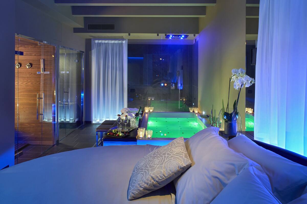 A modern hotel room with a large bed, a glass-enclosed jacuzzi, and a view of the night sky.