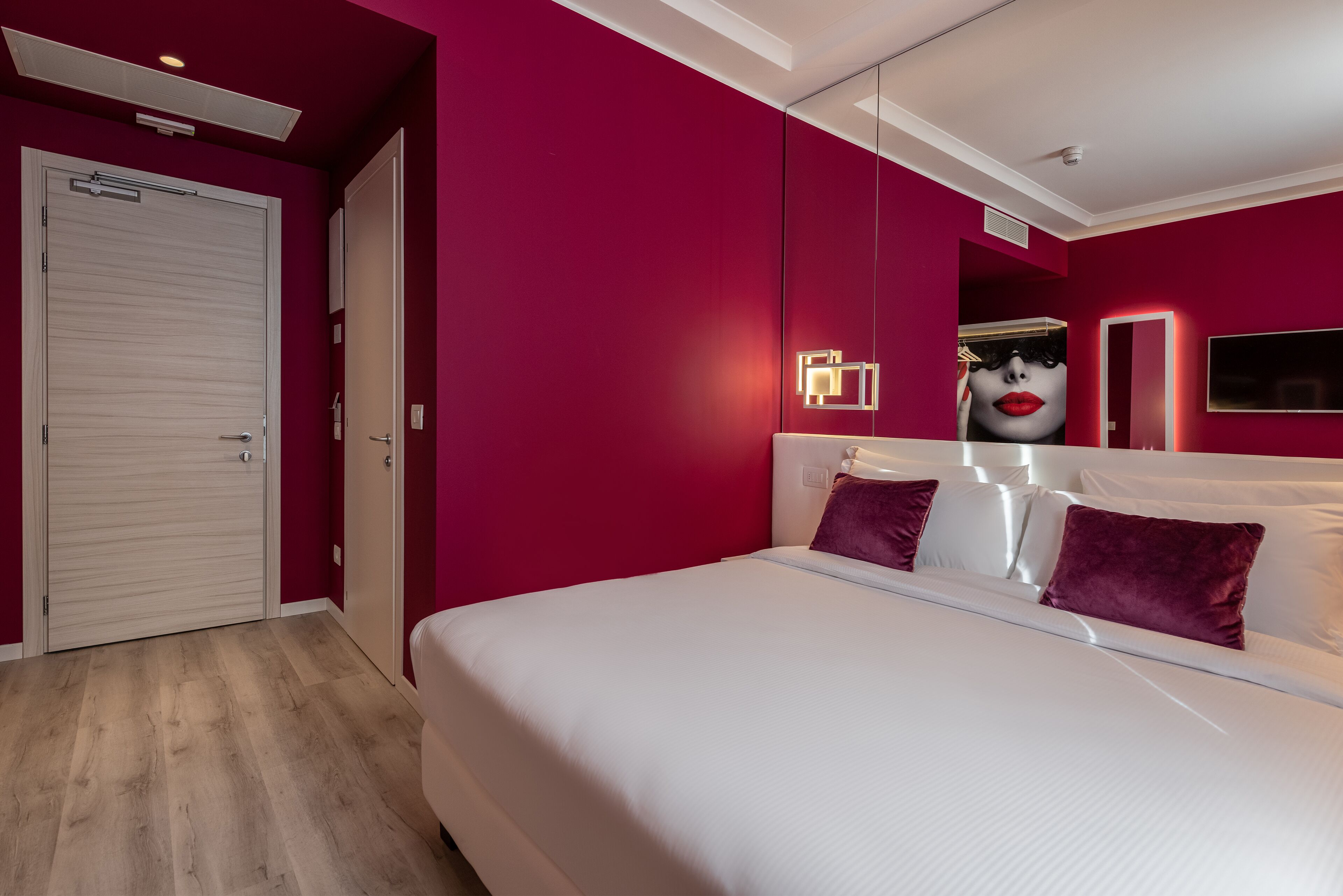 A modern bedroom with a large bed, a mirror, and a red accent wall.