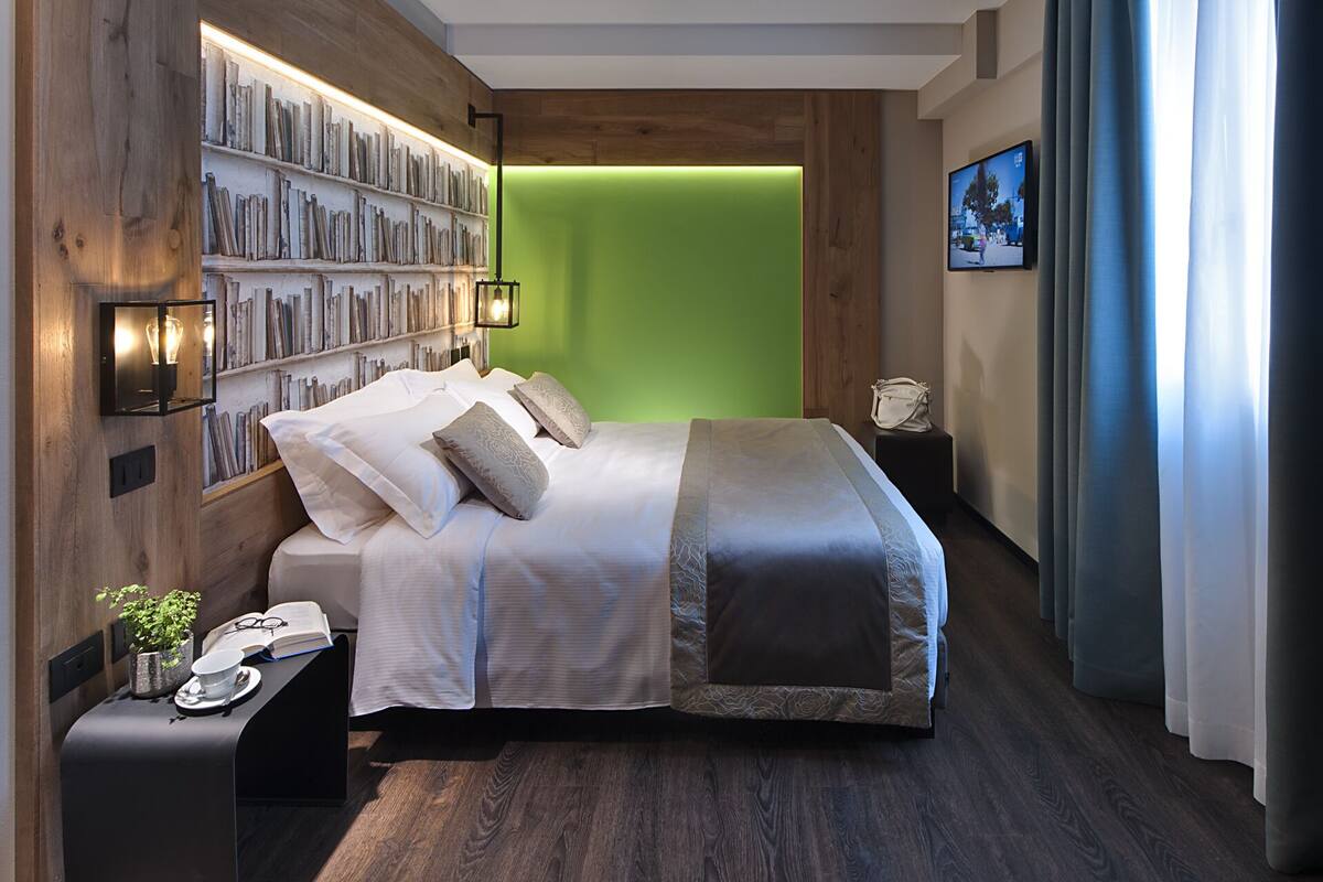 A modern hotel room with a large bed, a bookshelf, a green accent wall, and a television on the wall.