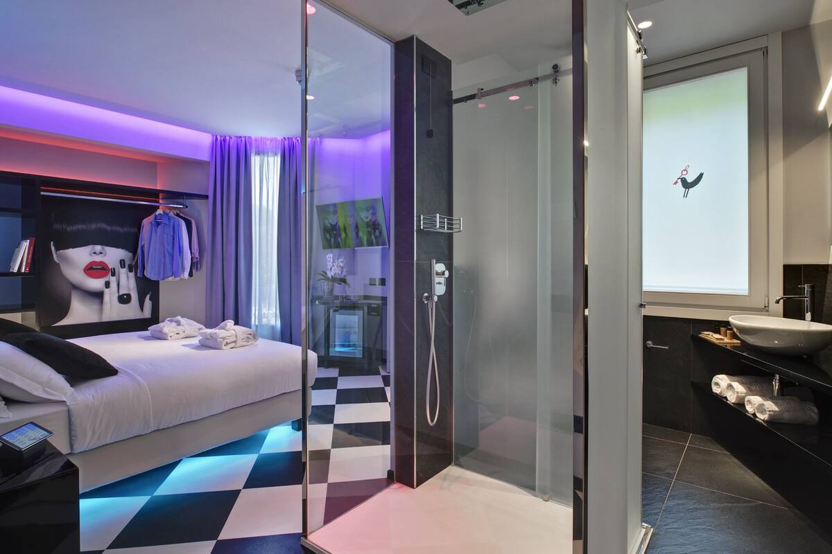 A modern hotel room with a checkered floor, a glass-enclosed shower, a bed with neatly folded towels, and a large window with a bird decoration.
