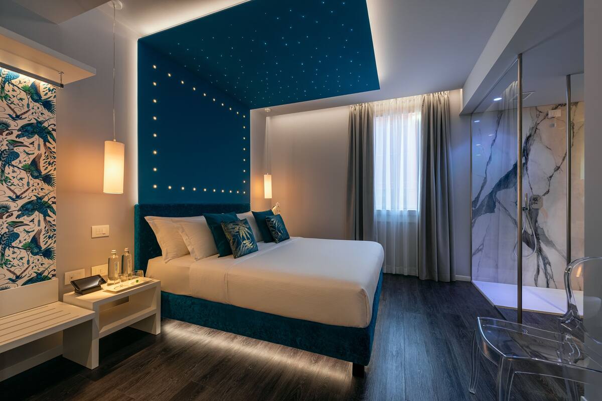 A modern hotel room with a large bed, a nightstand, a desk, and a glass shower area.