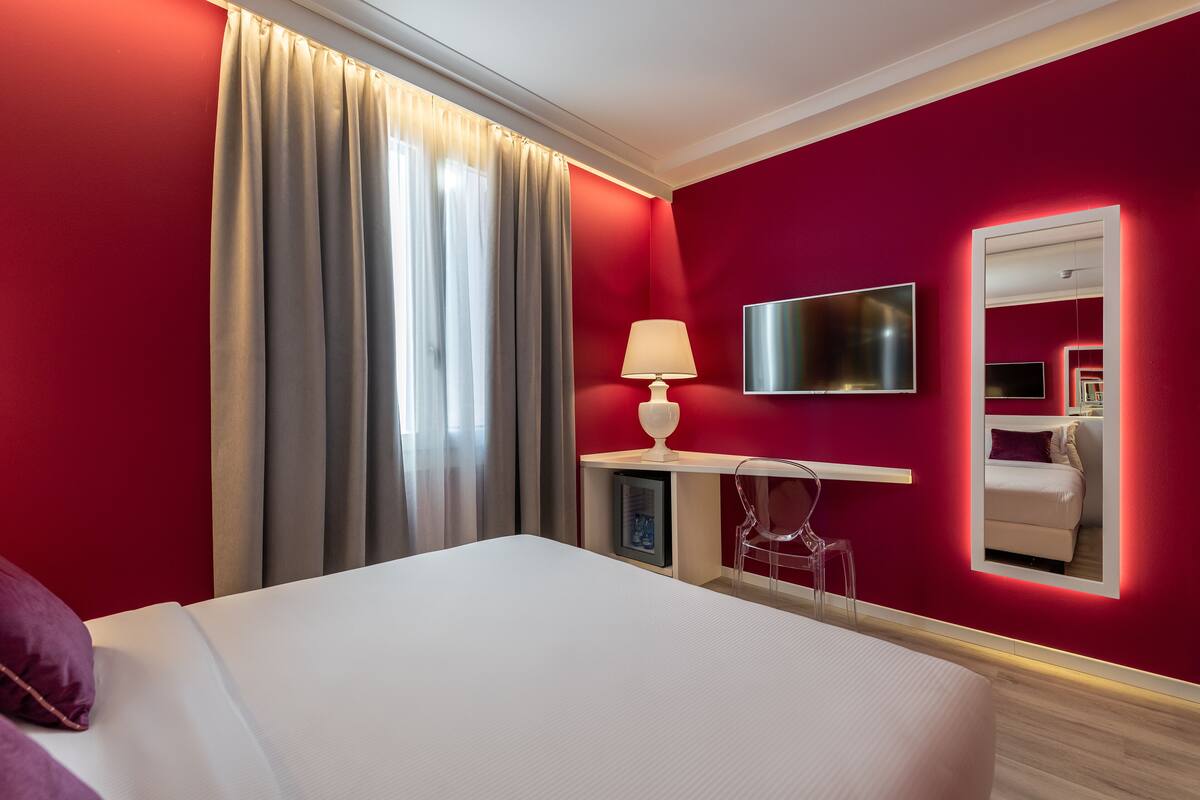 A hotel room with a large bed, a TV, a desk with a lamp, a chair, and a mirror.