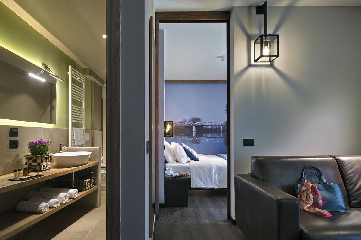 A modern bathroom with a large sink, a mirror, and a towel rack. There is a wooden shelf with towels and a basket of flowers.