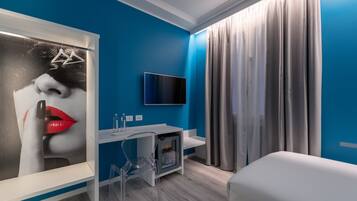 Una habitación de hotel moderna con una pared azul de acento, un televisor de pantalla plana sobre una consola blanca y un pequeño refrigerador, además de un sofá blanco.