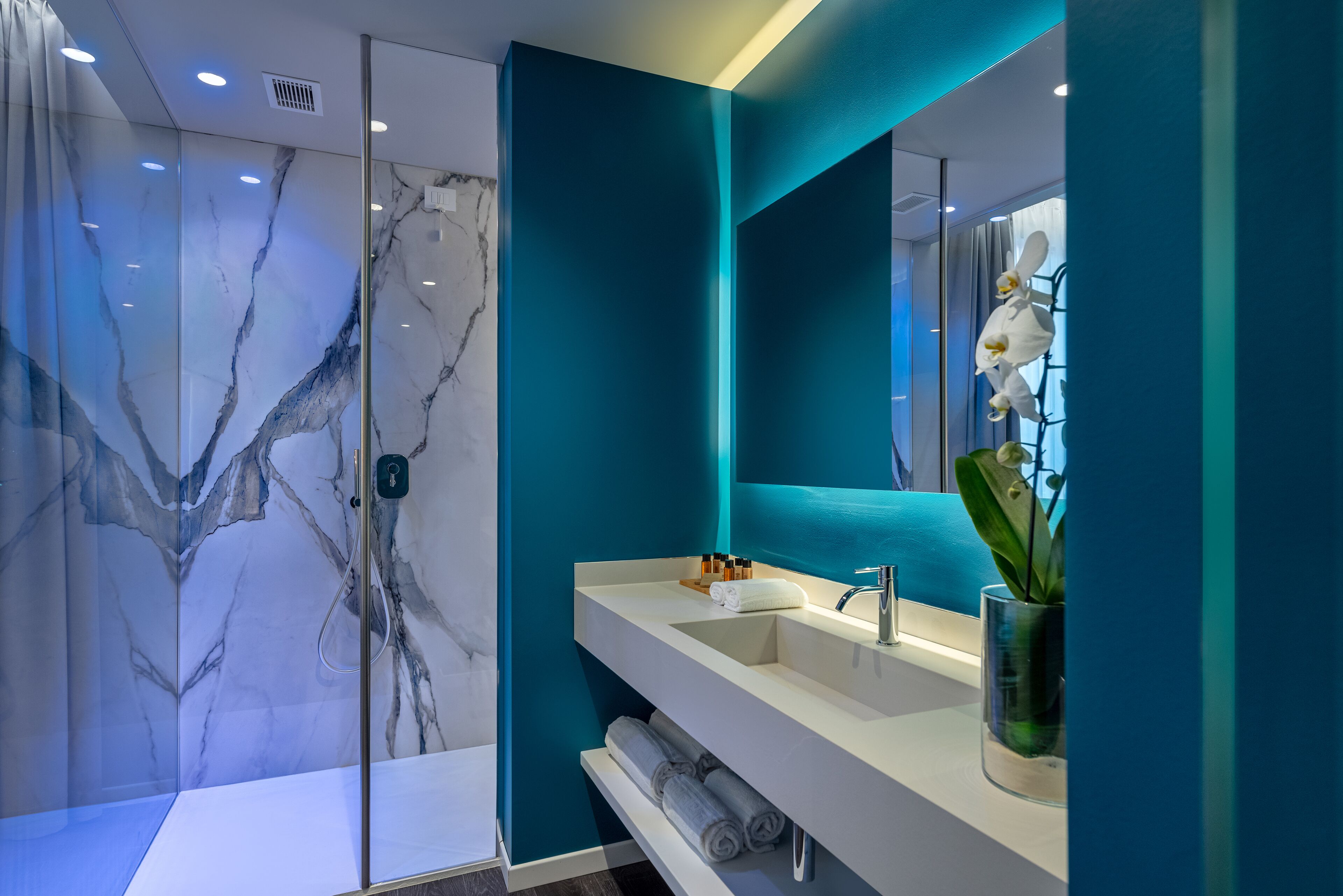 Une salle de bain moderne avec un mur en marbre, un mur d’accent de couleur turquoise, un lavabo blanc et un miroir.