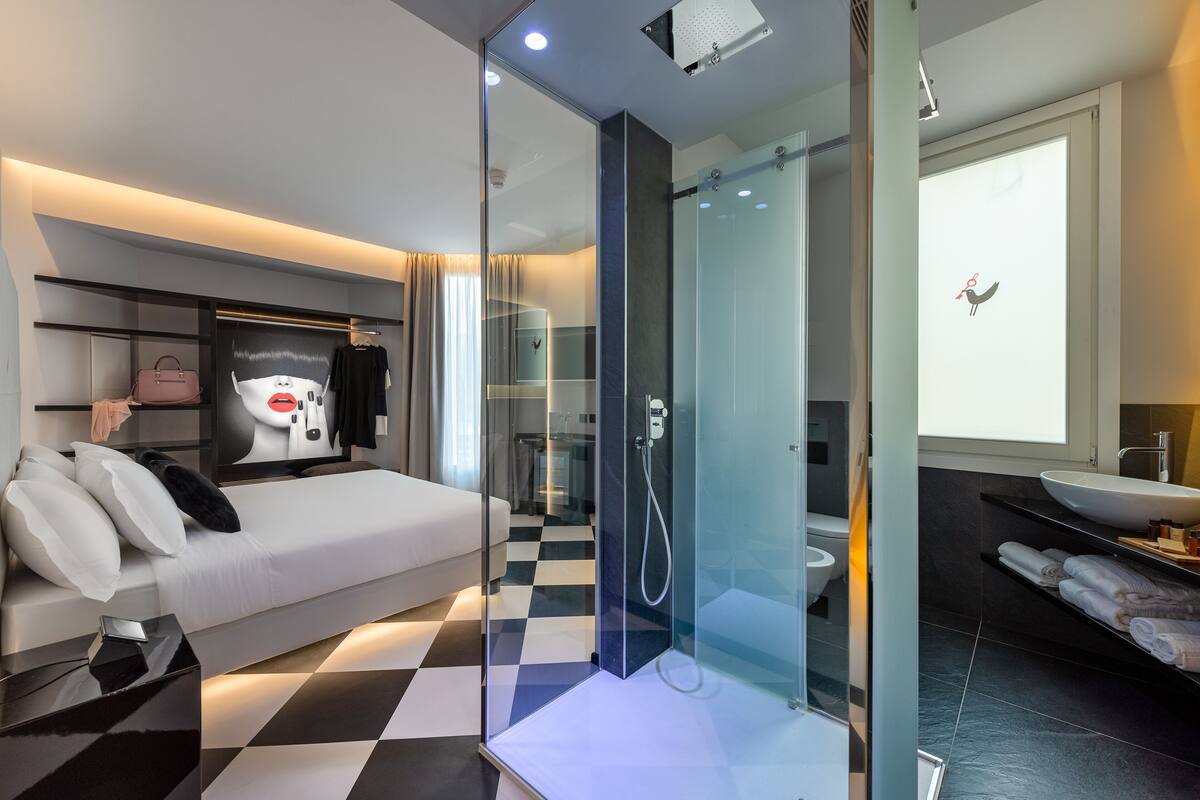 A modern hotel room with a checkered floor, a bed with white linens, a glass-enclosed shower, a large mirror, and a wall-mounted TV.