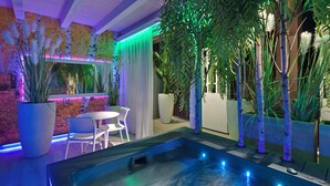 Un espace piscine intérieur moderne, doté d’un jacuzzi, entouré de plantes en pot et comprenant un coin salon avec une table et des chaises.