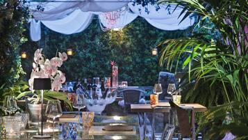 An outdoor dining area with tables set for a meal, surrounded by lush greenery and decorative lighting.