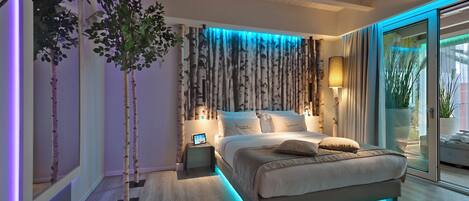 Un dormitorio moderno con una cama, mesitas de noche, un televisor montado en la pared y un ventanal con cortinas.
