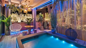 Un espace piscine intérieur moderne, doté d’un plancher en bois, d’un coin salon avec une chaise et une table, ainsi que de diverses plantes et éléments décoratifs.