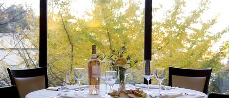 Una mesa redonda para dos personas, con una botella de vino, un florero con una sola flor y un cuenco de pan.