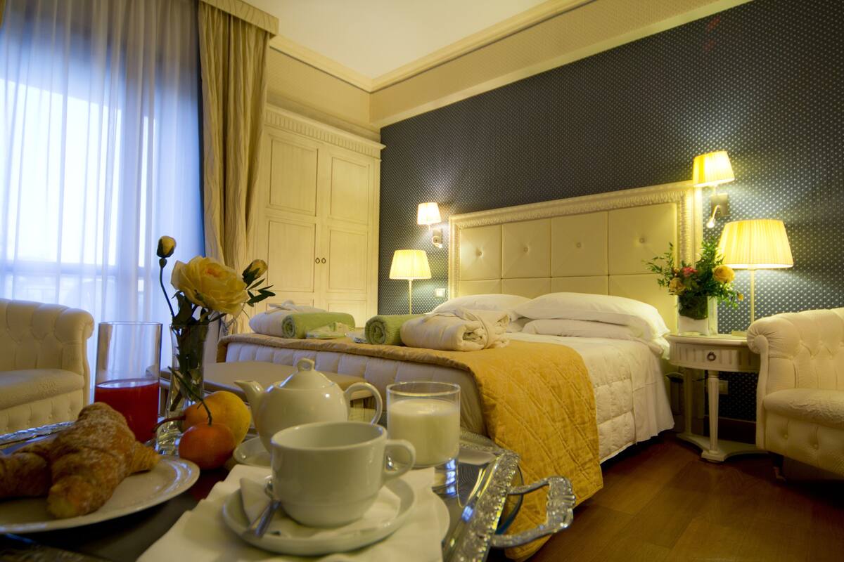 Hotelkamer met een bed, nachtkastjes, een zithoek met een stoel, en een ontbijttafel met croissants, fruit en drankjes.