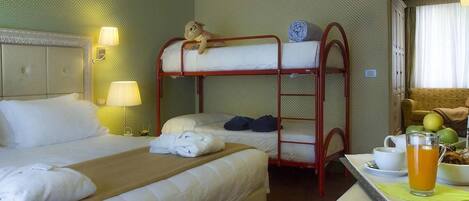 A hotel room with two beds, a bunk bed, a lamp, a table with a cup and a glass of orange juice, and a fruit bowl.