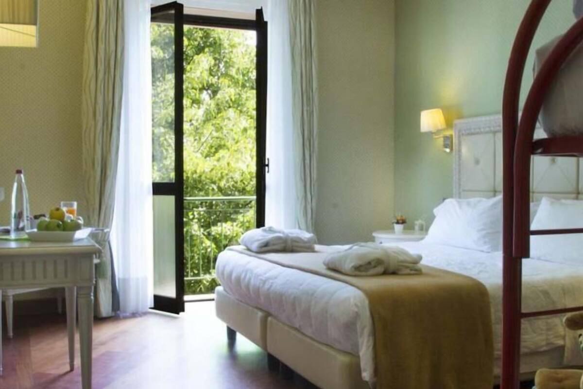 Hotelkamer met een bed, nachtkastje met lamp, stoel en uitzicht op groen door een geopende deur.
