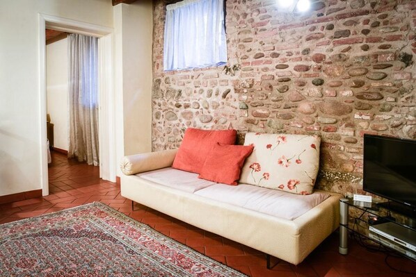 A cozy living room with a stone wall, a beige sofa with floral cushions, a red throw pillow, a patterned rug, and a television on a stand. - Residence Antico San Zeno (Verona)
