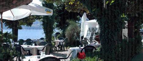 An outdoor dining area with tables and chairs, surrounded by greenery and overlooking a body of water.