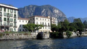 A waterfront building with balconies, a stone wall, and a mountain in the background. - Hotel Riviera (Griante)