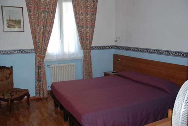 Quarto com cama de madeira, uma cadeira, radiador e janela com cortinas florais.