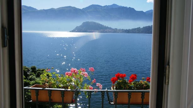 A view of a lake with mountains in the background, a balcony with potted plants, and a clear sky. - Hotel Riviera (Griante)