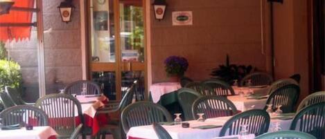An outdoor dining area with tables and chairs, a covered section with a red awning, and a building entrance with a clock and decorative elements.