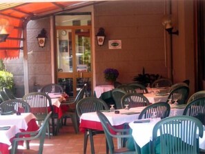 An outdoor dining area with tables and chairs, a covered section with a red awning, and a building entrance with a clock and decorative elements.