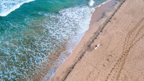 Vista aérea de una playa con olas, una persona con una tabla de surf y una orilla arenosa.
