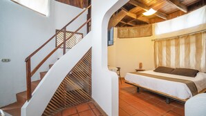 Un dormitorio con una cama, una mesita pequeña y una silla. Se ve una escalera con barandillas de madera que sube a un nivel superior.