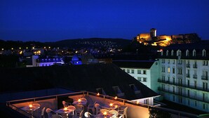 Eine Dachterrasse mit Tischen und Stühlen, mit Blick auf eine Stadtlandschaft, deren Hintergrund von einem markanten, schlossähnlichen Bauwerk dominiert wird.