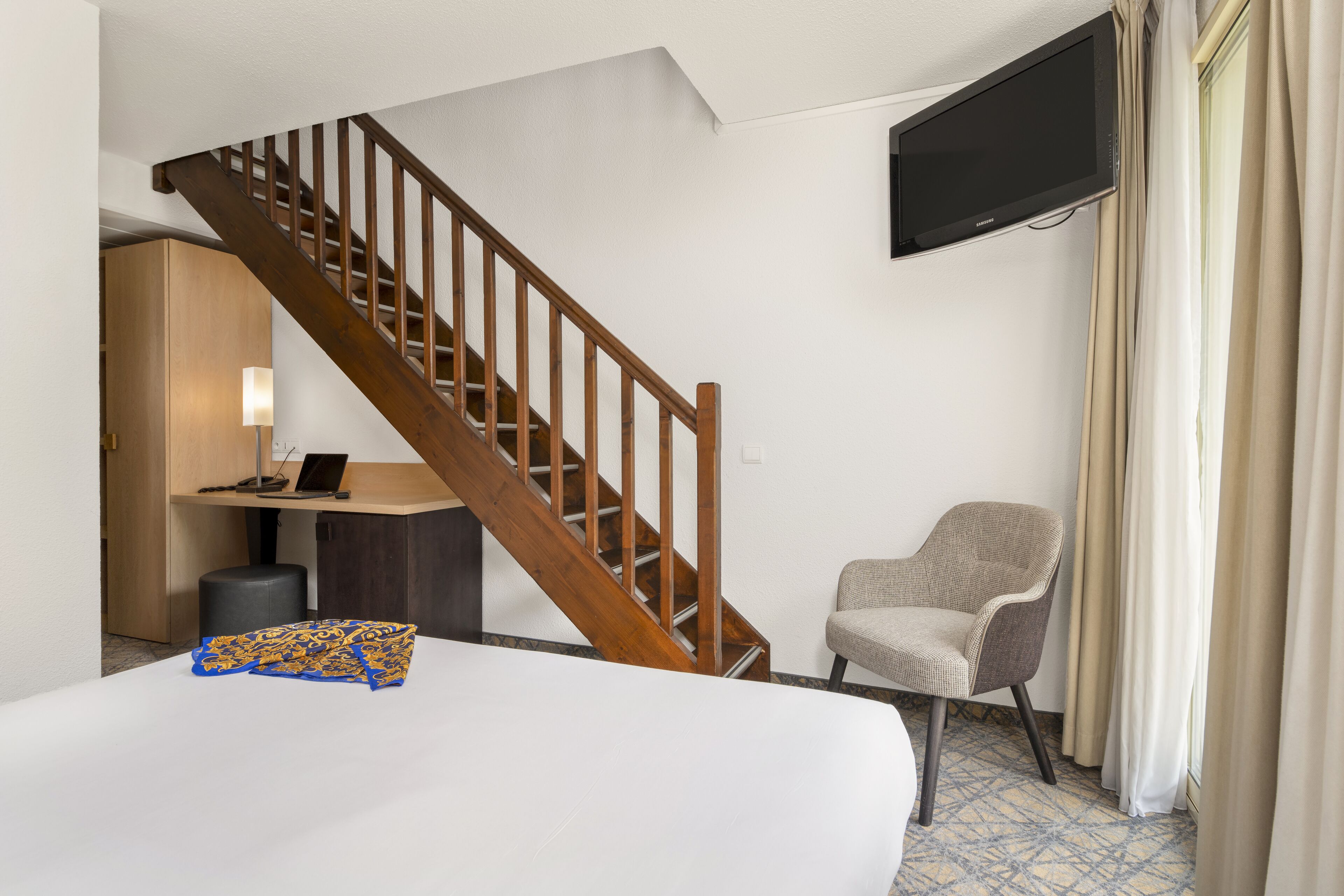 Een hotelkamer met een houten trap, een flatscreen televisie aan de muur, een bureau met een lamp, een stoel en een bed.
