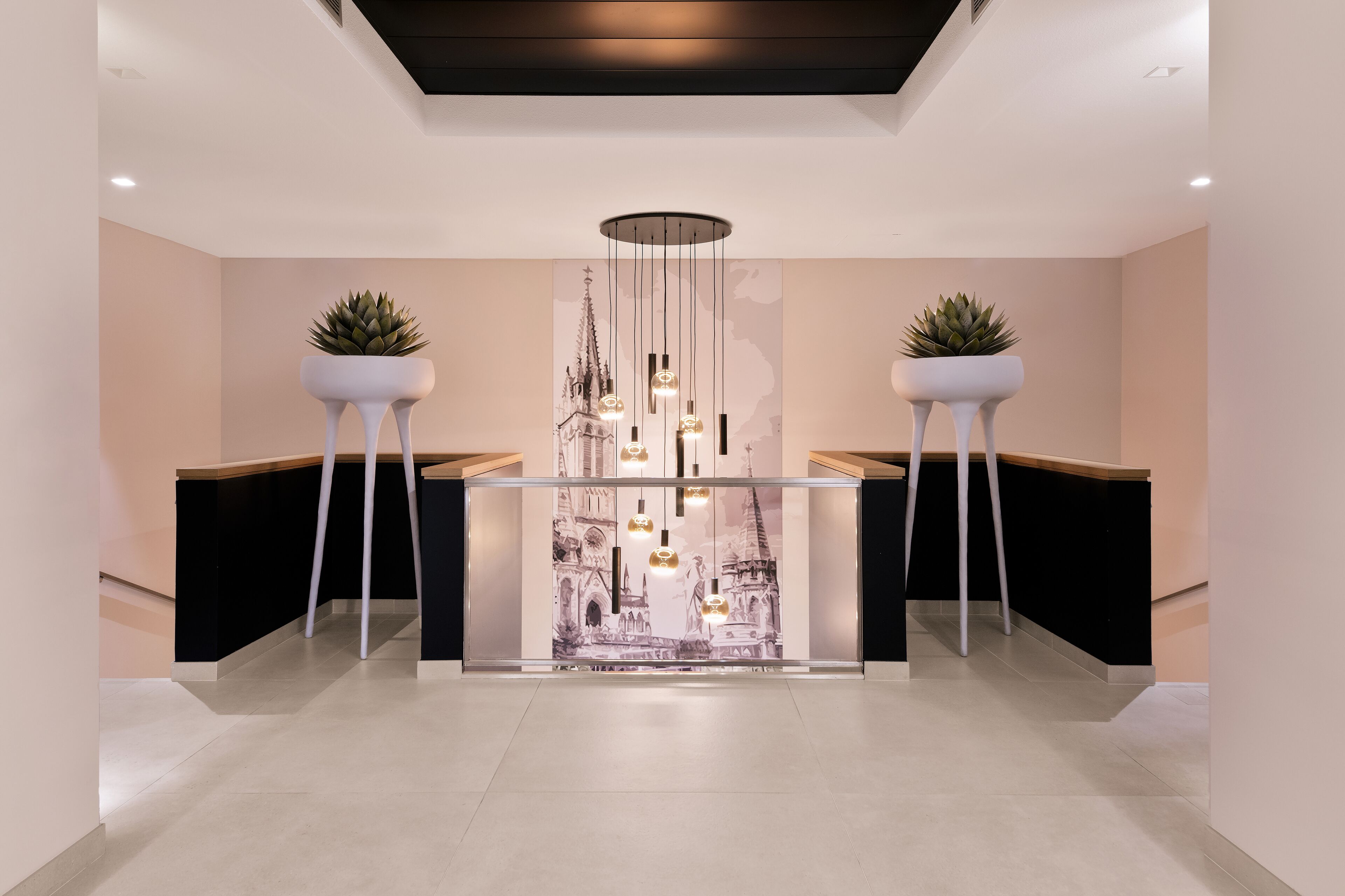 Een moderne receptiebalie met een glazen scheiding, twee potplanten, een kroonluchter en een muurschildering van een kathedraal.