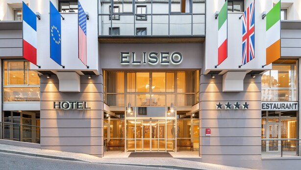 Das Elisseo Hotel von außen, mit Glastür, Flaggen und einem Schild, das auf ein Restaurant hinweist.