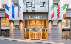 The exterior of the Elisseo Hotel with a glass entrance, flags, and a sign indicating a restaurant inside. - Hotel Eliseo (Lourdes)
