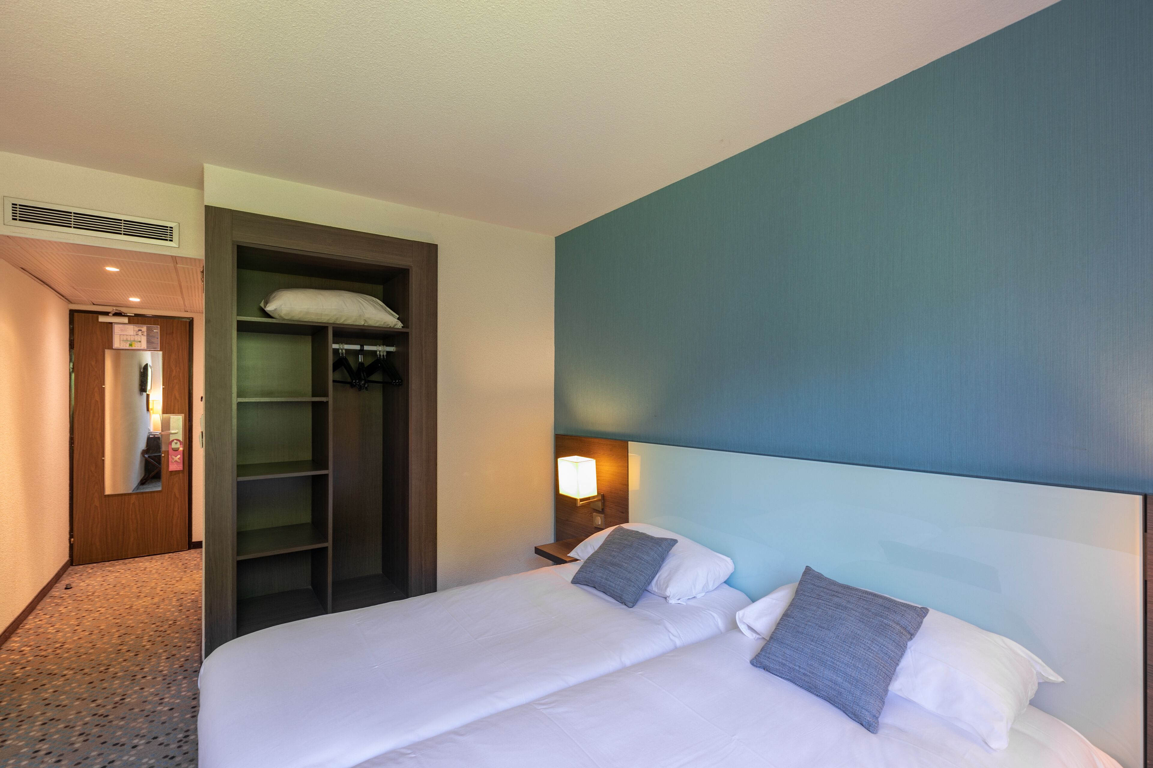 Habitación de hotel con una cama grande, un armario, una mesita de noche con lámpara y una puerta que da a un baño.