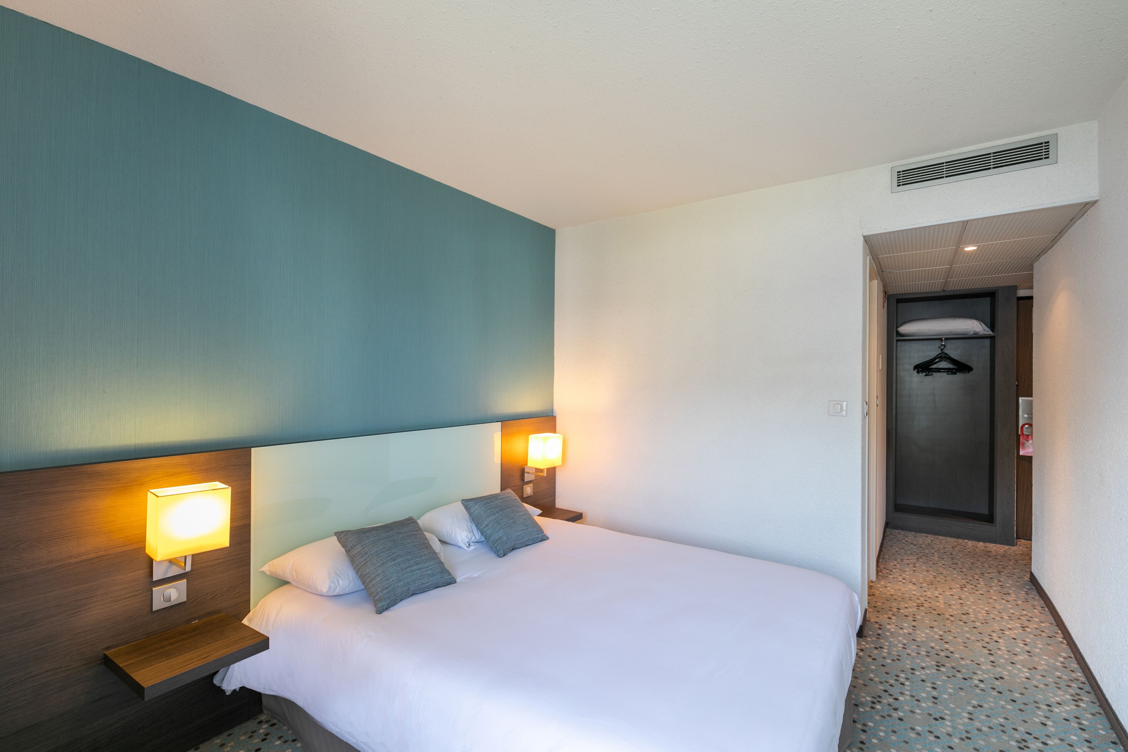 A hotel room with a neatly made bed, two bedside lamps, and a door leading to a hallway.