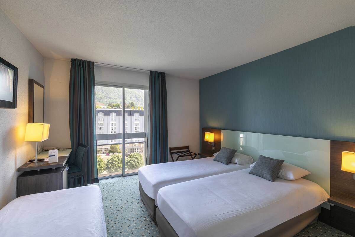 A hotel room with two beds, a desk, a chair, and a large window offering a view of buildings and trees.
